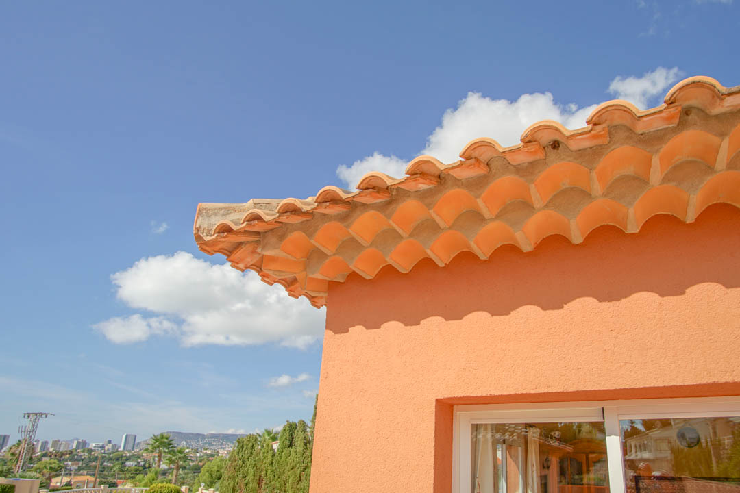Detail of the traditional terracotta tile roof and decorative eaves against a peach facade, offering distant views of the city skyline and mountains.