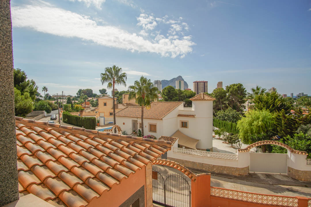 Elevated view from the upper level showcasing traditional terracotta roofing, lush palm trees, and a clear vista of the Peñón de Ifach on the horizon.