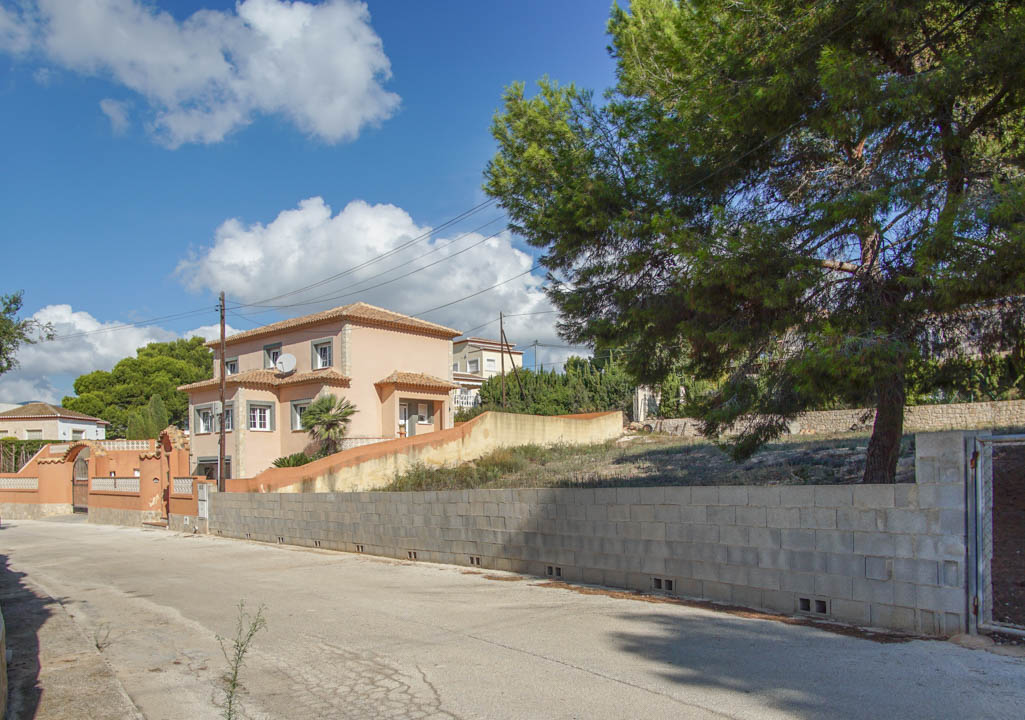 Exterior view of the three-story villa from the street, featuring the perimeter wall, tiled roof, and mature pine trees bordering the property.