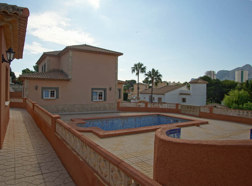 Exterior view of the villa featuring a spacious stone-paved terrace, a blue mosaic-tiled pool, and multi-level architecture with traditional clay roof tiles.