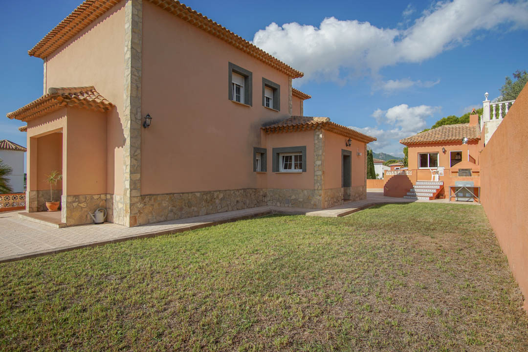 Exterior view of the villa featuring earth-toned walls, natural stone accents, and a lawn. The background shows the separate outdoor kitchen and wood-fired oven.