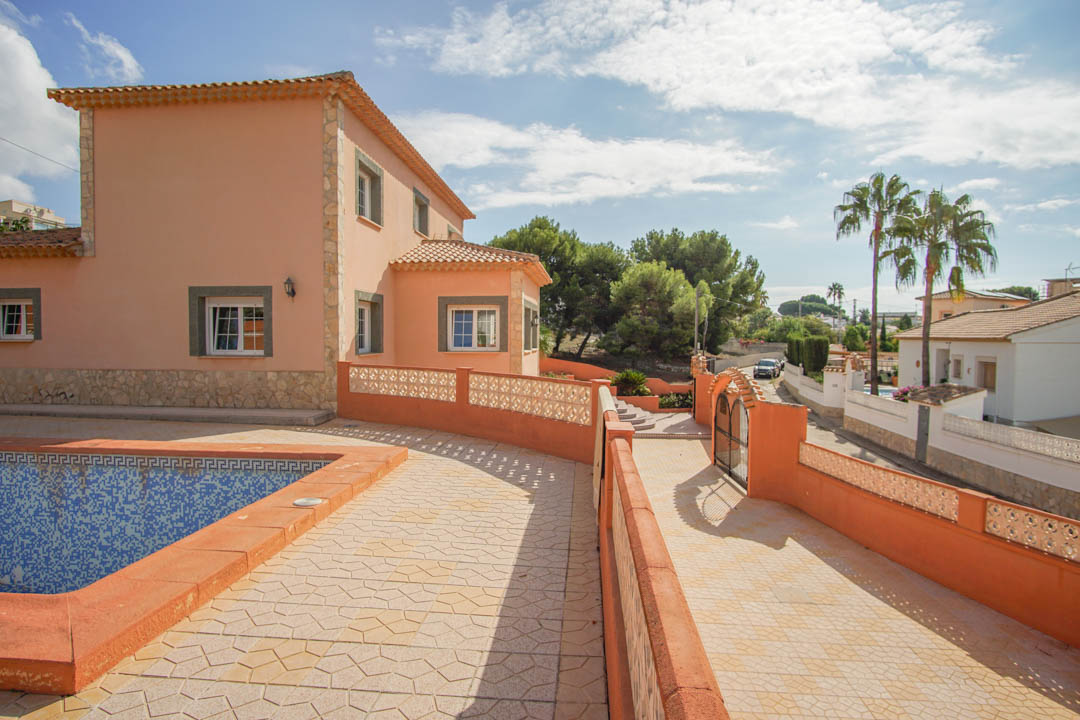 Exterior view of the villa featuring the swimming pool area, paved driveway leading to the gate, and the facade with stone accents and terracotta roof tiles.