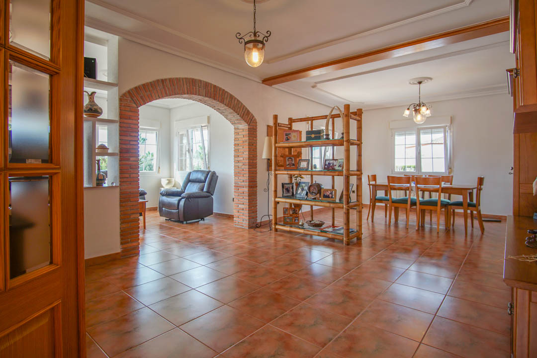 Living and dining area featuring a prominent exposed brick archway, terracotta-style floor tiles, and solid wood cabinetry. Natural light fills the space through multiple windows.