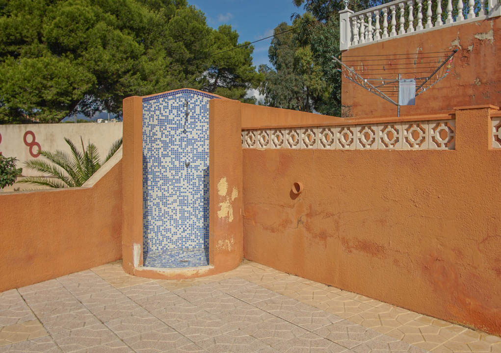 Outdoor shower featuring blue and white mosaic tiling, set against terracotta-toned walls with decorative breeze blocks and patterned stone paving.