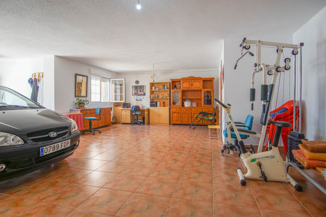 Spacious multi-purpose ground floor area featuring ceramic tiling, currently utilized as a garage and home gym with solid wood cabinetry and office workstations.
