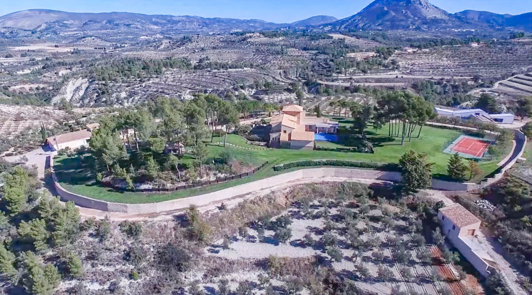 Aerial view of the estate featuring the main villa, swimming pool, and tennis court, set within landscaped grounds and surrounded by mountain scenery.