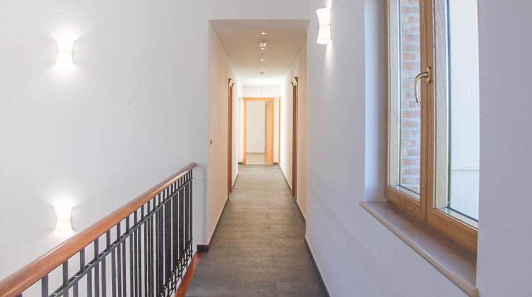 Bright corridor featuring grey stone flooring, high-quality wooden carpentry, and a staircase railing with metal balusters and a wooden handrail.