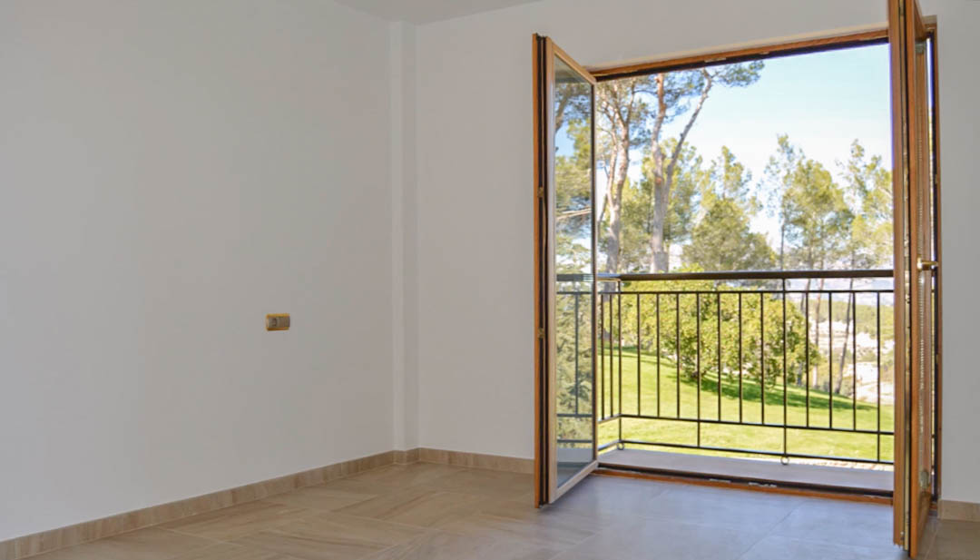 Bright room featuring tiled flooring and high-quality wooden double doors opening onto a private balcony with views of the surrounding pine trees and lawn.