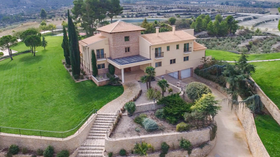 Elevated view of the main villa featuring stone masonry, terracotta roofing, and landscaped gardens with stone walls, set against a backdrop of terraced agricultural land.