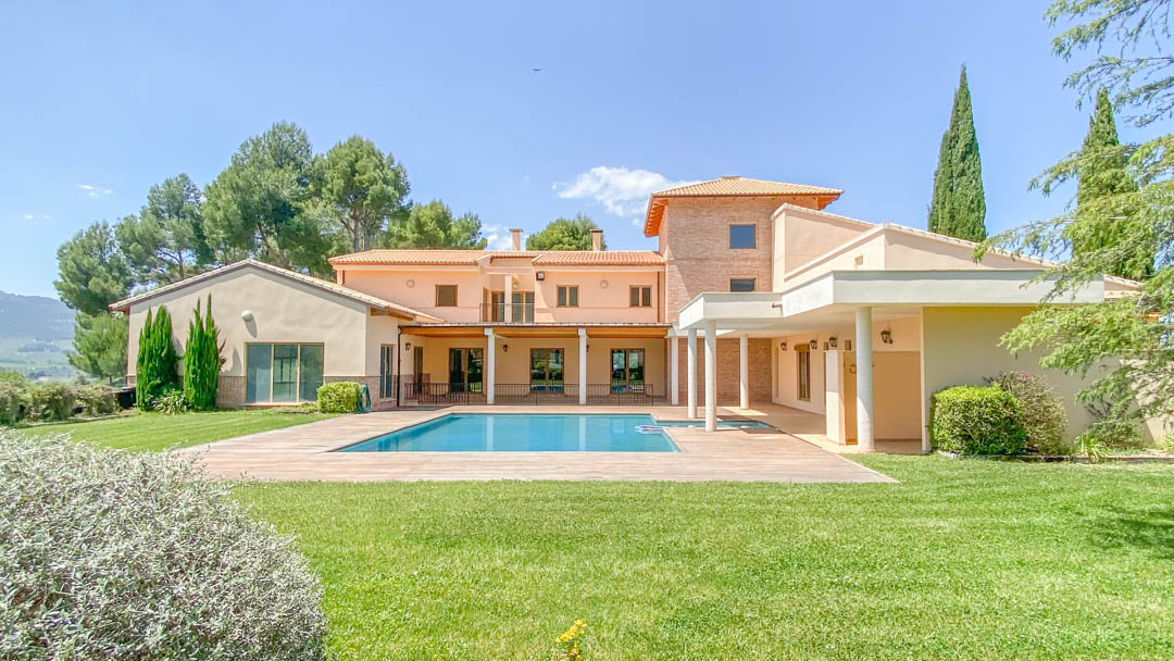 Exterior view of the main villa featuring a rectangular swimming pool, expansive lawn, and covered porches with white columns against a mountain backdrop.
