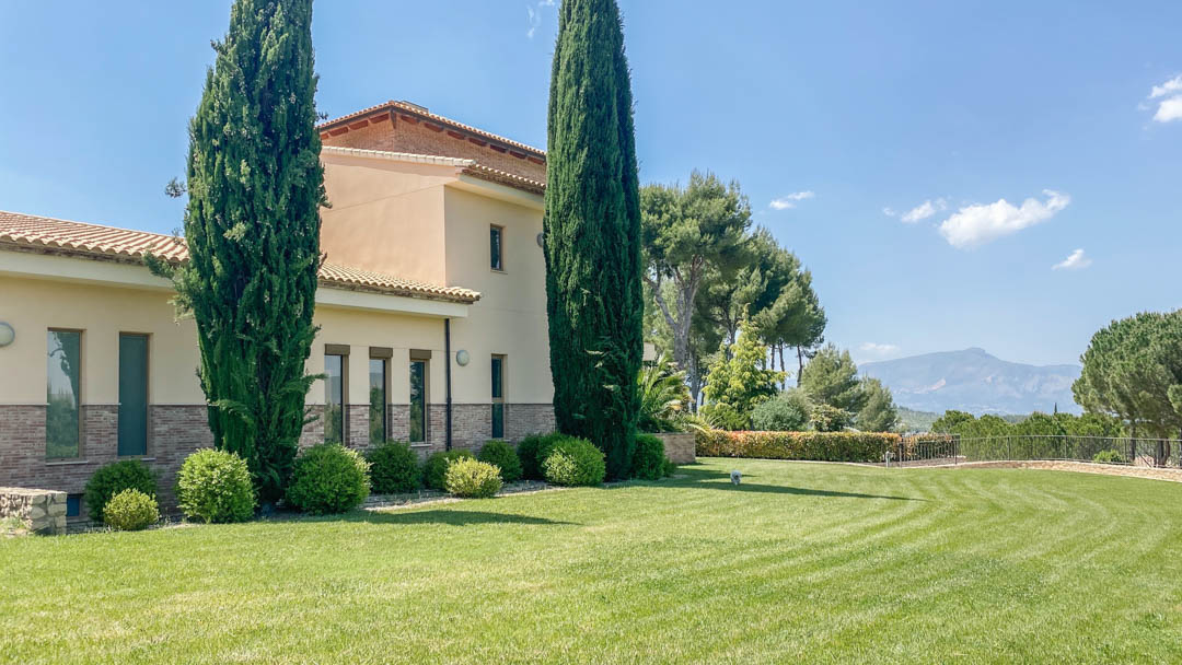 Exterior view of the main villa featuring stone-clad walls, manicured lawns, and tall cypress trees against a backdrop of distant mountains.