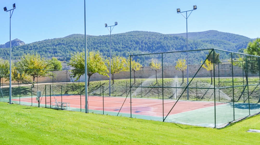 Fenced tennis court featuring a red and green surface, professional floodlighting for night matches, and panoramic views of the surrounding mountains.