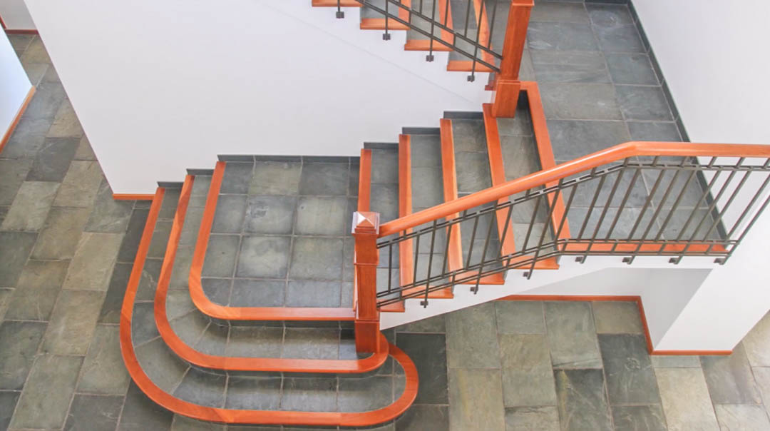 Grand staircase featuring natural stone tiling with polished wood trim. The design includes a dark metal railing with a matching wooden handrail against minimalist white walls.