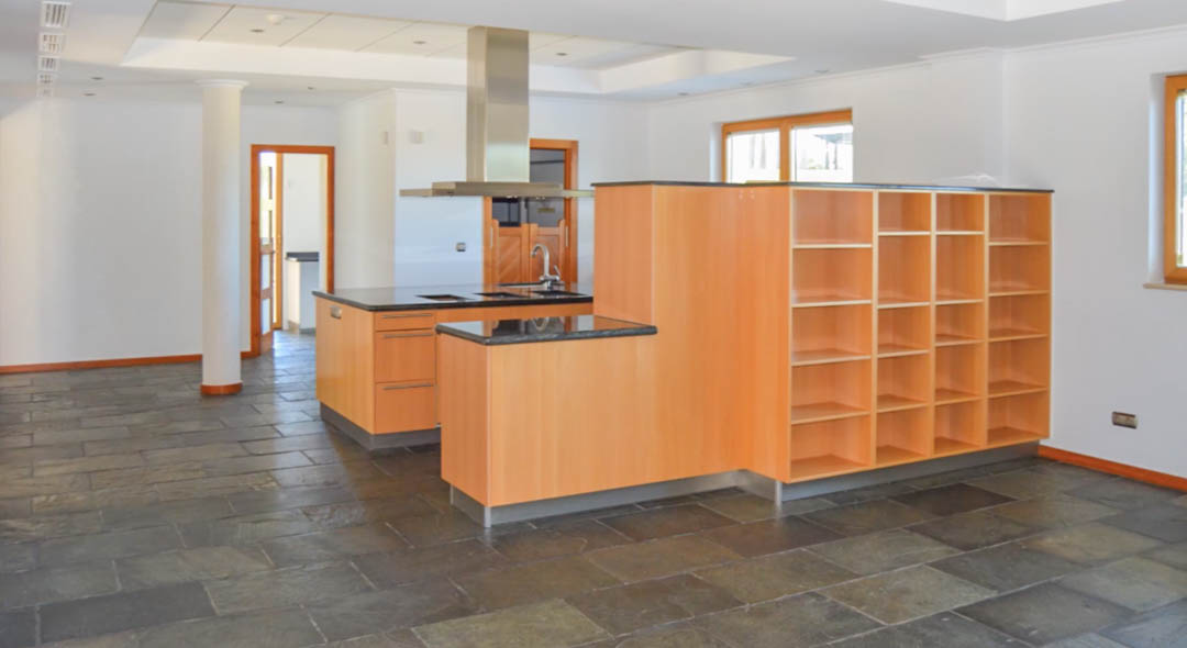 Open-concept kitchen featuring a central island with dark granite countertops, natural stone flooring, light wood cabinetry, and integrated shelving under recessed lighting.