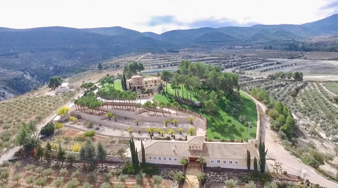 Panoramic aerial view showcasing the main villa and annex building, surrounded by terraced gardens with palms, pines, and vast agricultural groves set against a mountain backdrop.