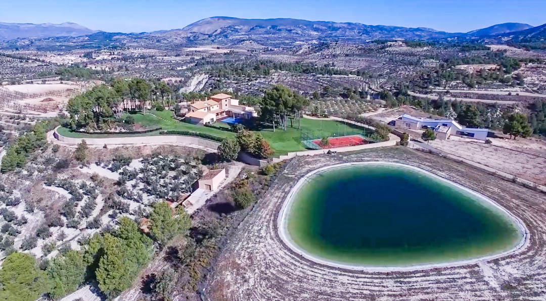 Panoramic aerial view showing the main villa, private water reservoir, and red tennis court, all set within a vast landscape of mountains and agricultural fields.