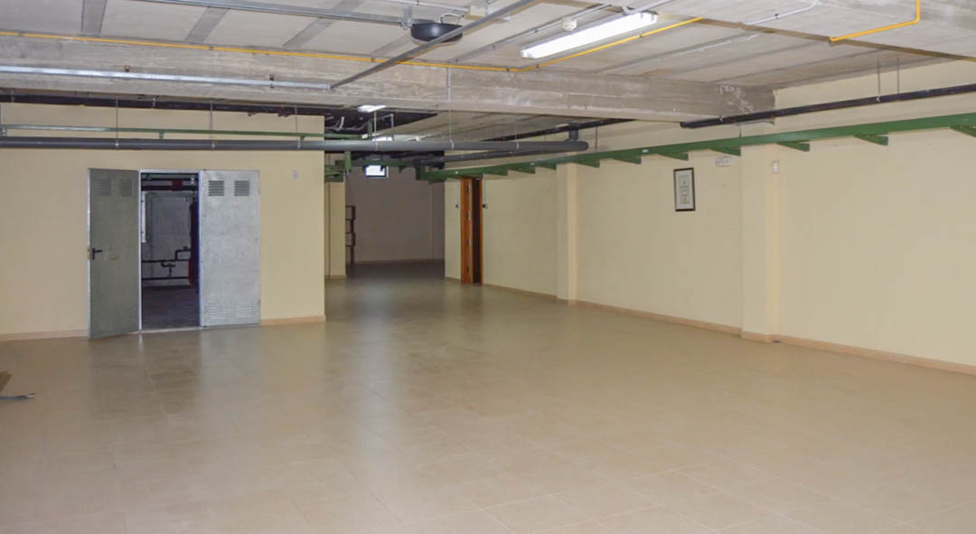 Spacious underground garage featuring tiled flooring, exposed concrete ceiling beams, and visible technical installations. Includes metal doors leading to maintenance areas.