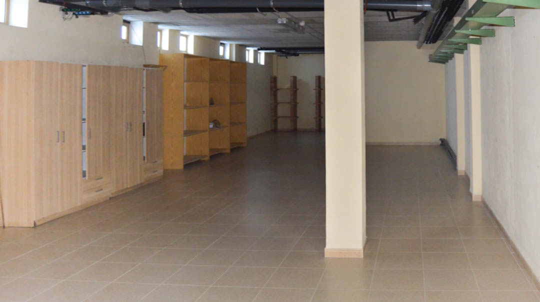 Spacious underground storage area featuring ceramic tiled flooring, wooden wardrobes, and open shelving units alongside exposed ceiling conduits.
