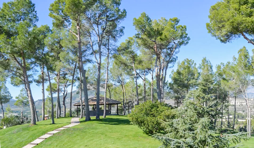 Tiled-roof gazebo set on a landscaped hillside, surrounded by mature pine trees with a stone path leading to panoramic mountain views.