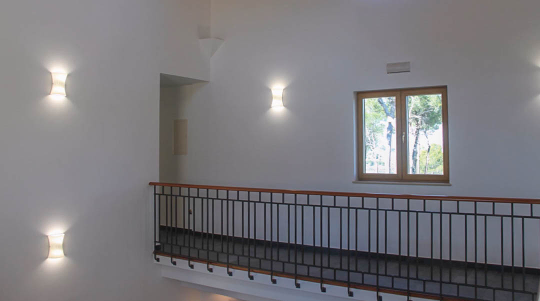 Upper landing featuring a wrought iron railing with wooden handrail, illuminated by modern wall sconces and a window overlooking the surrounding trees.