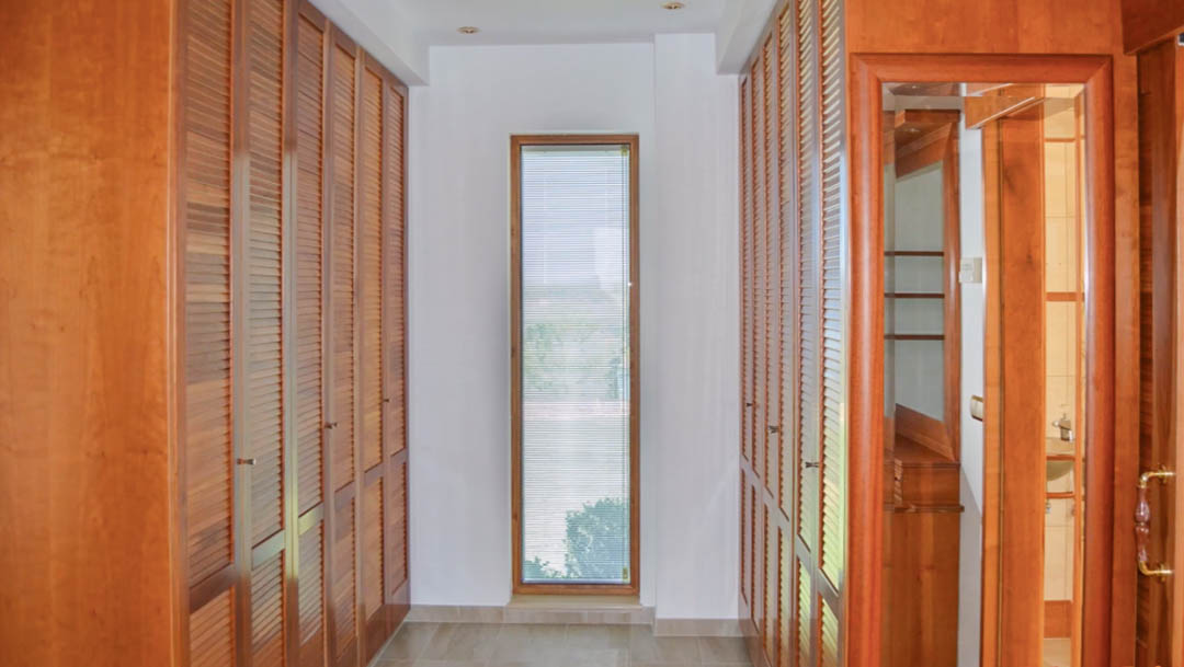 Walk-in closet featuring floor-to-ceiling wooden wardrobes with louvred doors, natural light from a narrow window, and recessed ceiling lighting.