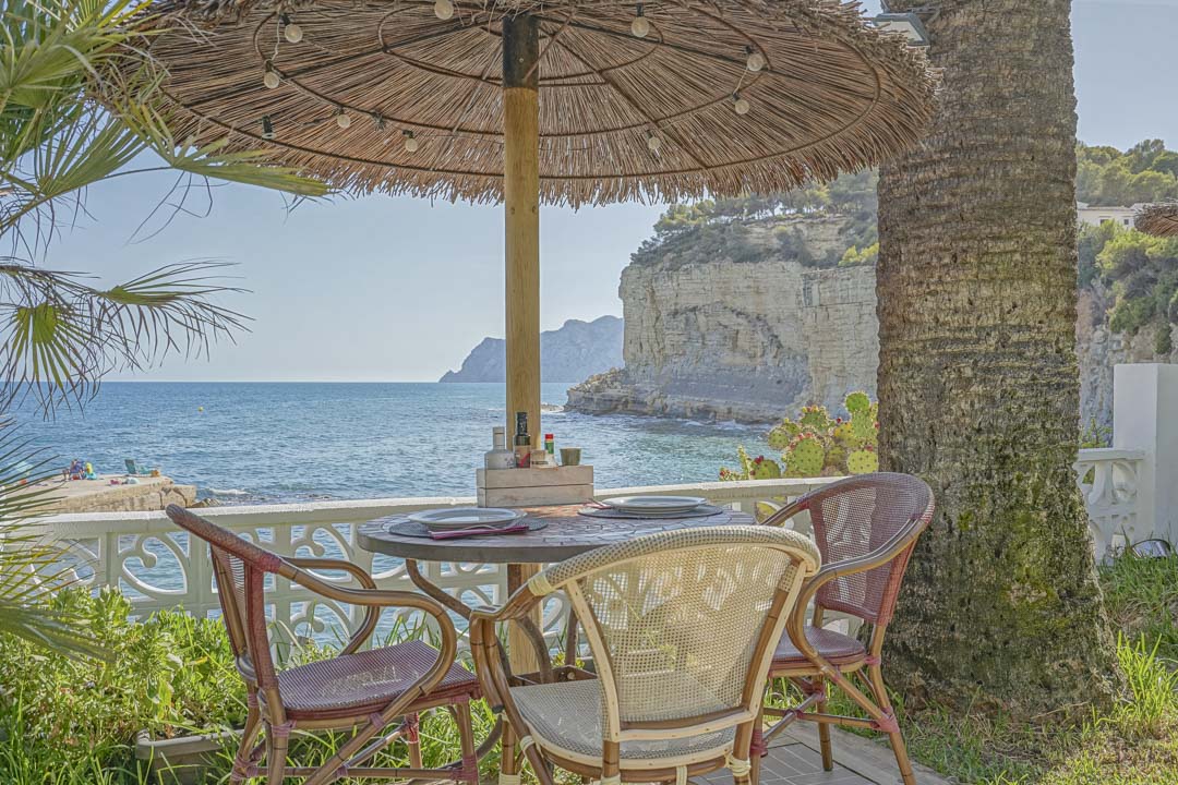 Outdoor dining area with a thatched umbrella and string lights, featuring bistro seating and direct views of the Mediterranean sea and coastal cliffs.