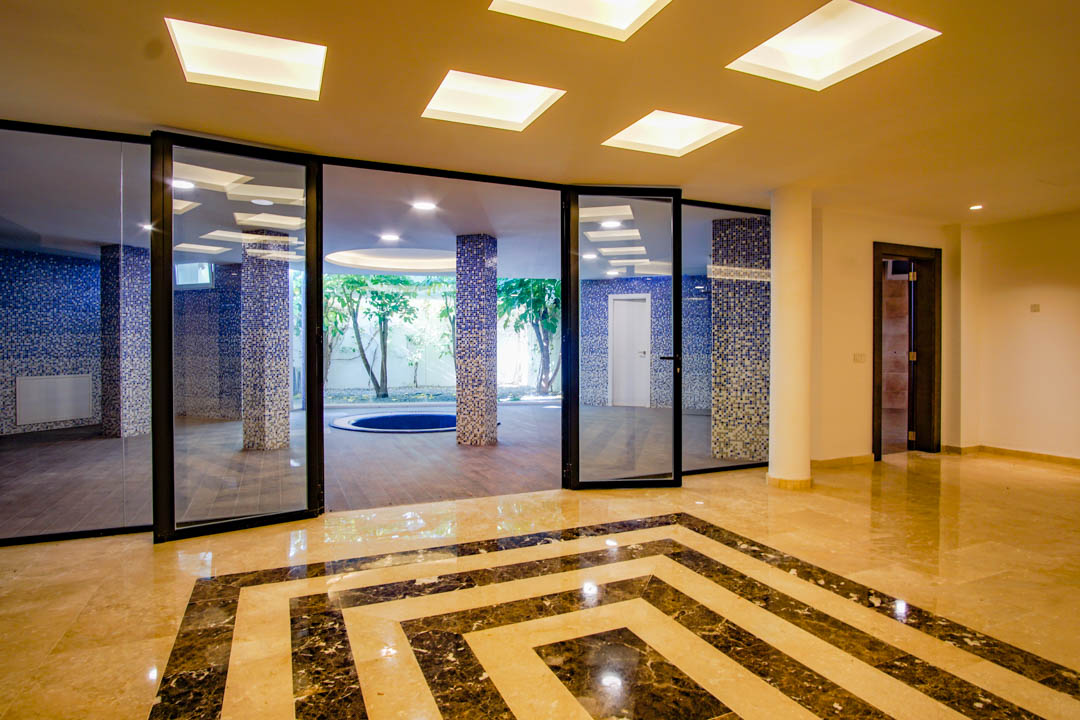 Basement wellness area featuring polished marble floors with geometric patterns, blue mosaic columns, and a view of the English patio with natural trees.