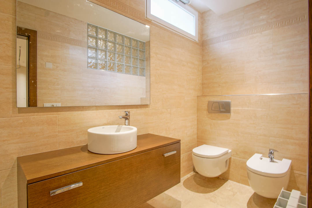 Bathroom featuring beige tiling, a floating wooden vanity with a vessel sink, wall-mounted toilet and bidet, and a large mirror reflecting a glass block partition.