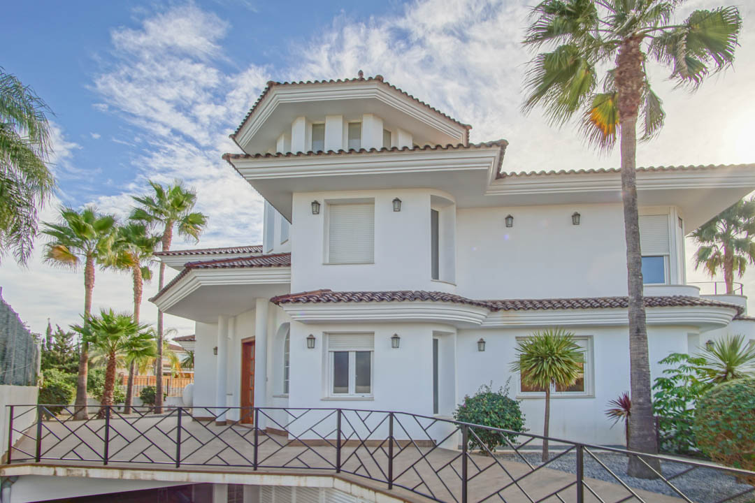 Colonial-style exterior featuring white facades, terracotta tile roofs, and black wrought-iron railings, framed by tall palm trees.