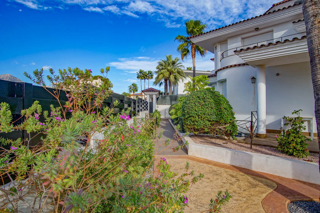 Colonial-style white facade with columns and porch, set within a mature garden featuring palm trees, flowering shrubs, and paved pathways under a clear sky.