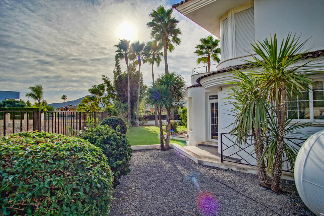 Colonial-style white facade with curved architectural lines, surrounded by a garden with tall palms, manicured shrubs, and gravel paths.