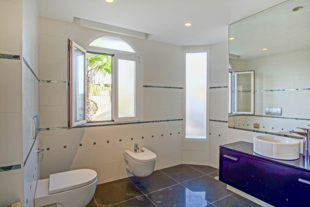 Contemporary bathroom featuring wall-mounted fixtures, a dark vanity with a vessel sink, and white wall tiles. An arched window brings in natural light and garden views.