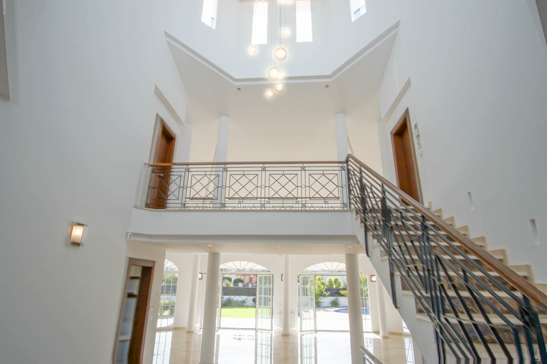 Double-height entrance hall featuring a marble staircase with wrought iron railings, an upper gallery with columns, and abundant natural light from arched garden doors.