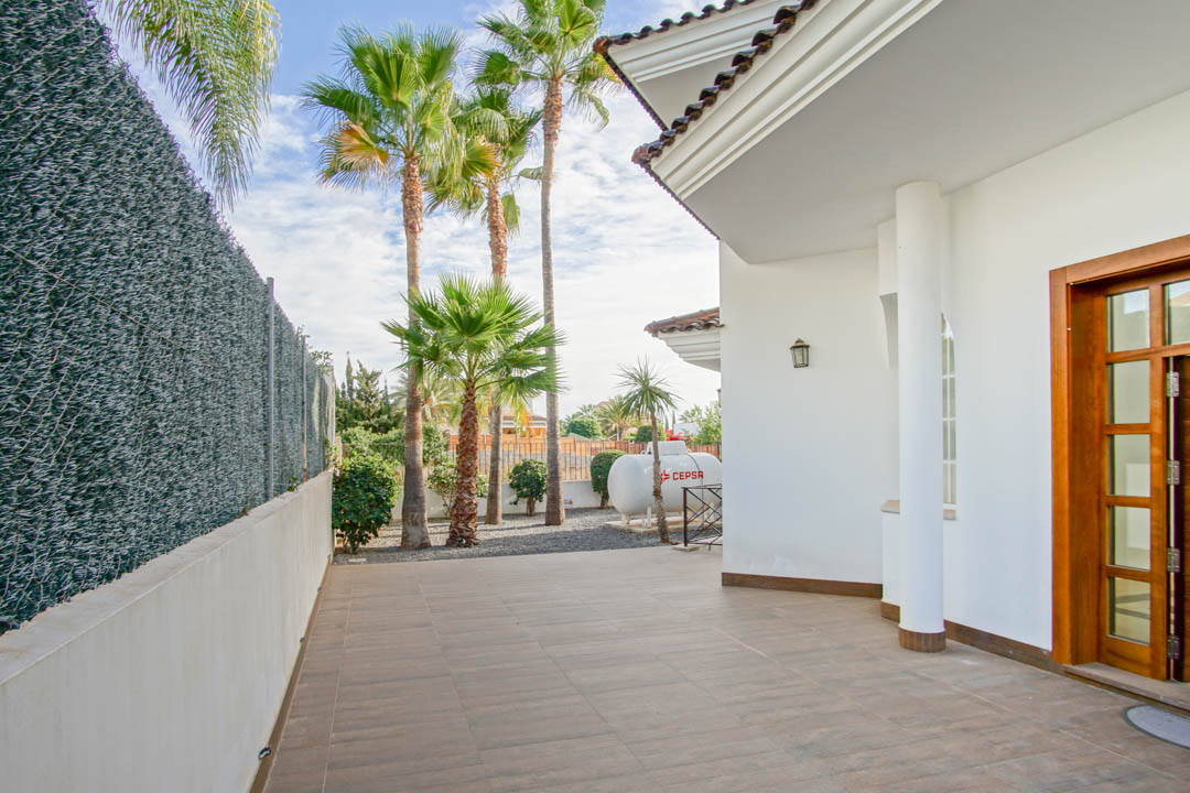 Exterior view featuring a tiled patio, white facade with a supporting column, wooden entrance door, and a garden area with mature palm trees.