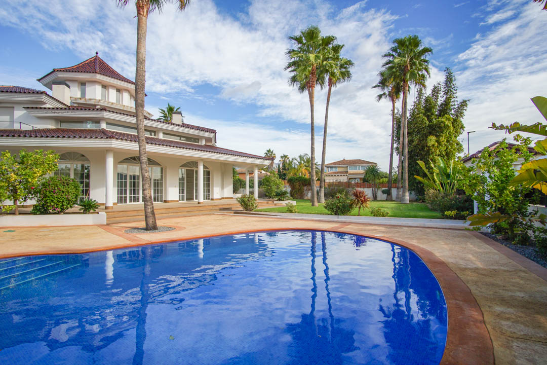 Exterior view of the colonial villa featuring a curved swimming pool, mature palm trees, and a white facade with arched windows and columns.