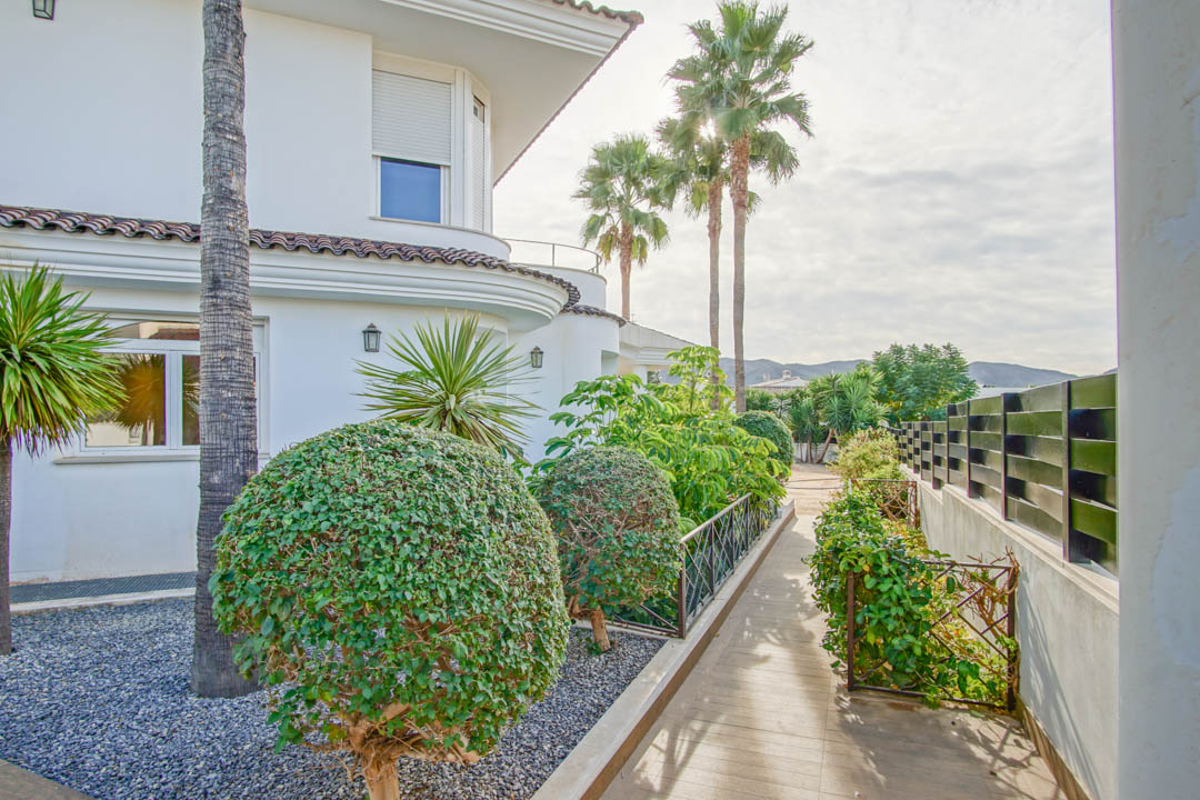 Exterior view of the side garden featuring paved pathways, manicured topiary bushes, and mature palm trees against the white colonial-style facade.