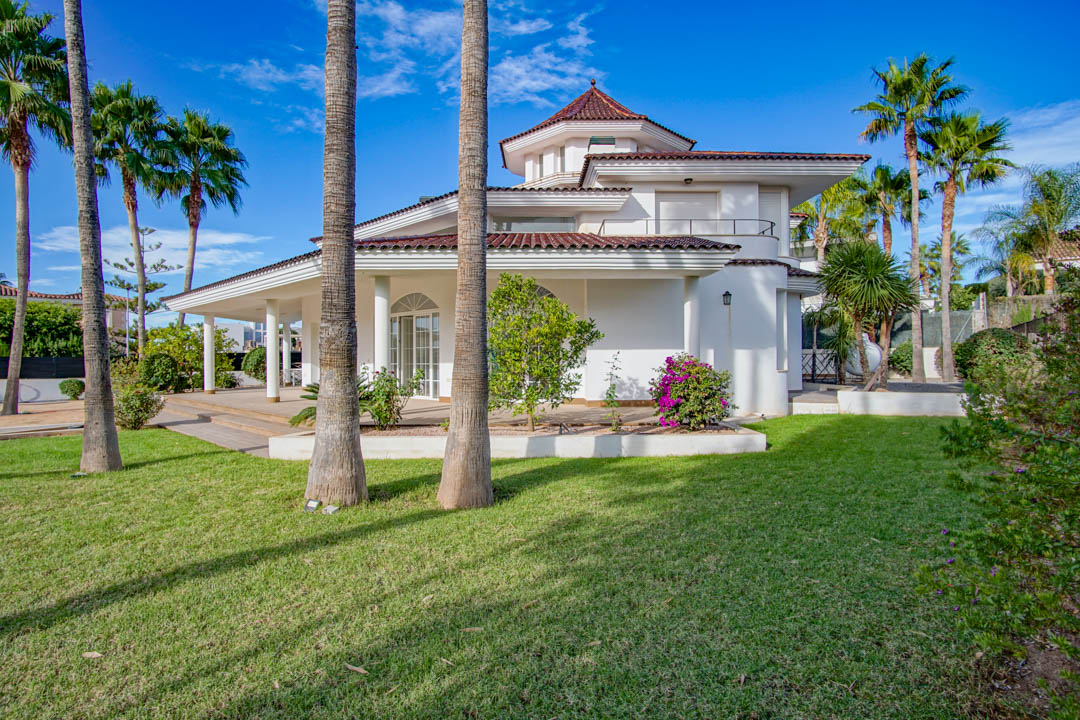Exterior view of the white colonial villa featuring a manicured lawn, tall palm trees, and traditional tiled roofing across multiple levels and terraces.