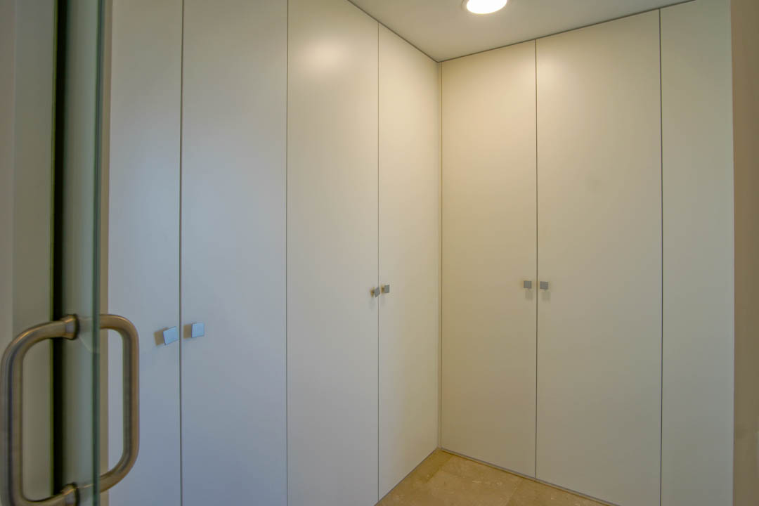Floor-to-ceiling built-in wardrobes in white with square metallic handles and natural stone flooring, illuminated by recessed ceiling lights.
