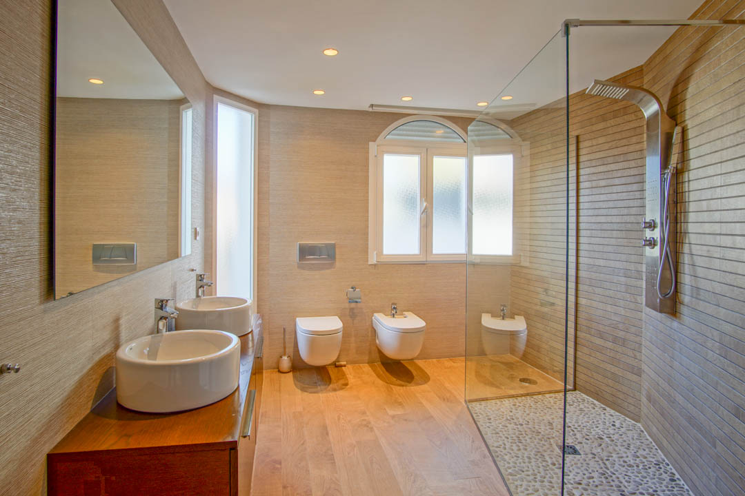 Modern bathroom featuring a double vessel sink vanity, a walk-in shower with pebble flooring, and wall-hung sanitary ware with warm recessed lighting.
