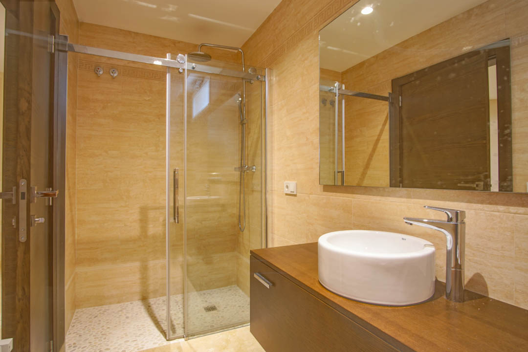 Modern bathroom featuring a glass-enclosed shower with pebble flooring, a circular vessel sink on a dark wood vanity, and warm beige tiling.