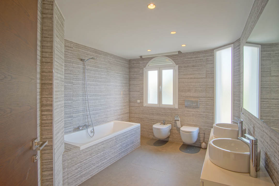 Modern bathroom featuring double vessel sinks, an integrated bathtub, and textured wall tiling. The space includes wall-hung sanitary ware and natural light from an arched window.