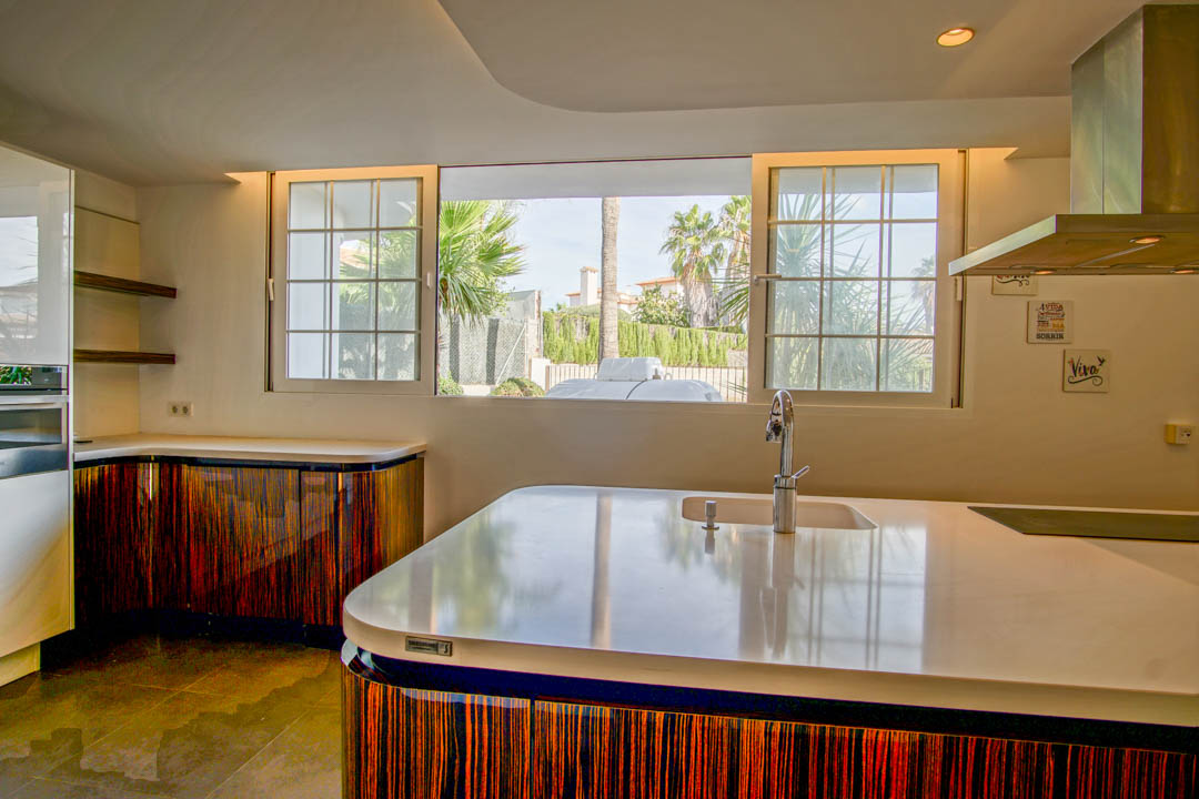 Modern kitchen featuring a white island, curved cabinetry with a dark wood finish, and large windows providing natural light and garden views.