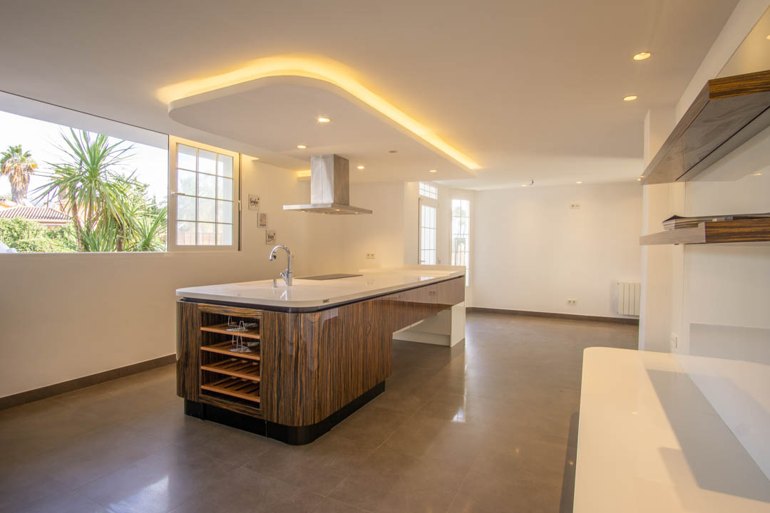 Modern kitchen featuring a wood-grain central island with white countertops and a stainless steel hood, complemented by indirect LED ceiling lighting and garden views.