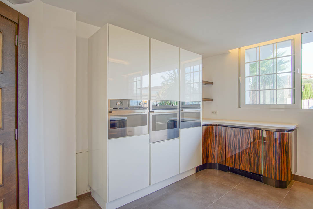 Modern kitchen featuring high-gloss white cabinetry, integrated stainless steel ovens, and curved lower units with a distinctive dark wood grain finish.