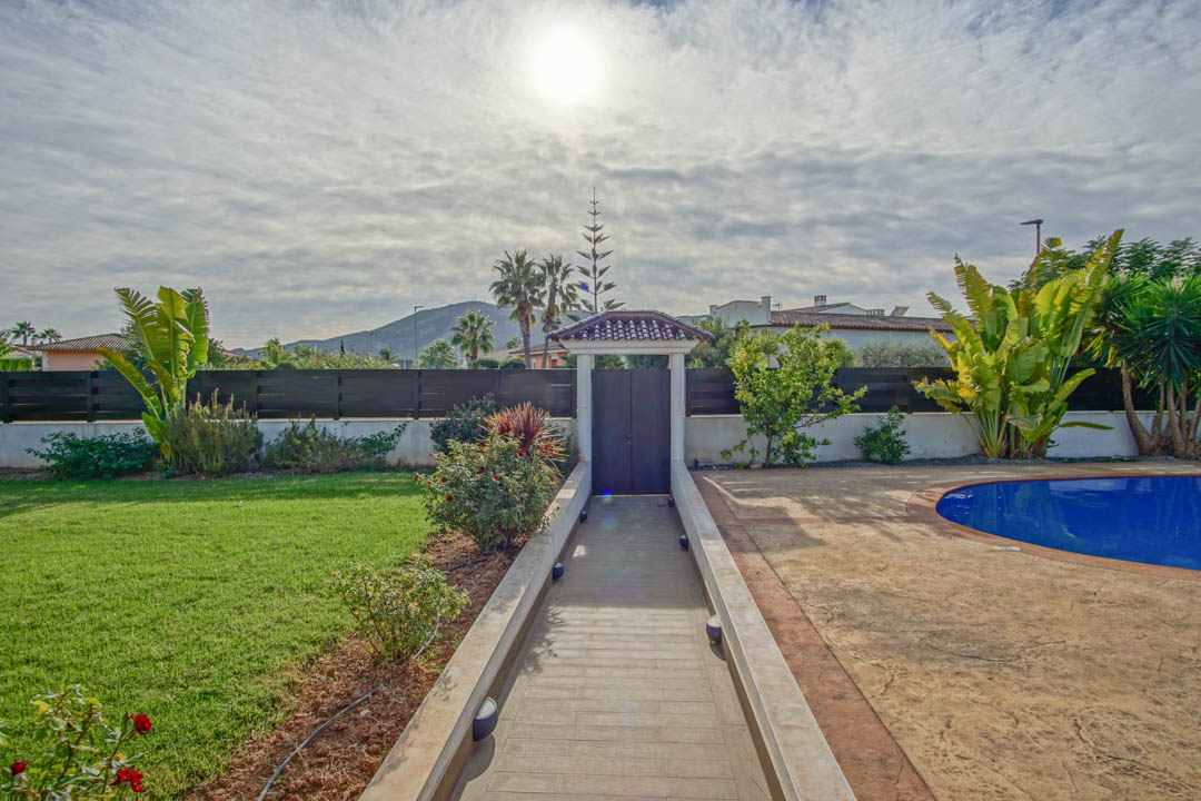 Paved walkway leading to a tiled entrance gate, bordered by a green lawn and a curved pool area with mature palms and mountain views.