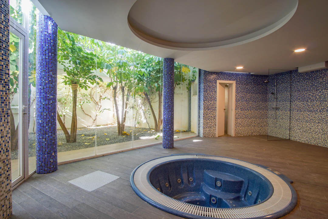 Spa area featuring a circular built-in jacuzzi, blue mosaic pillars, and floor-to-ceiling glass walls overlooking a private English courtyard with trees.