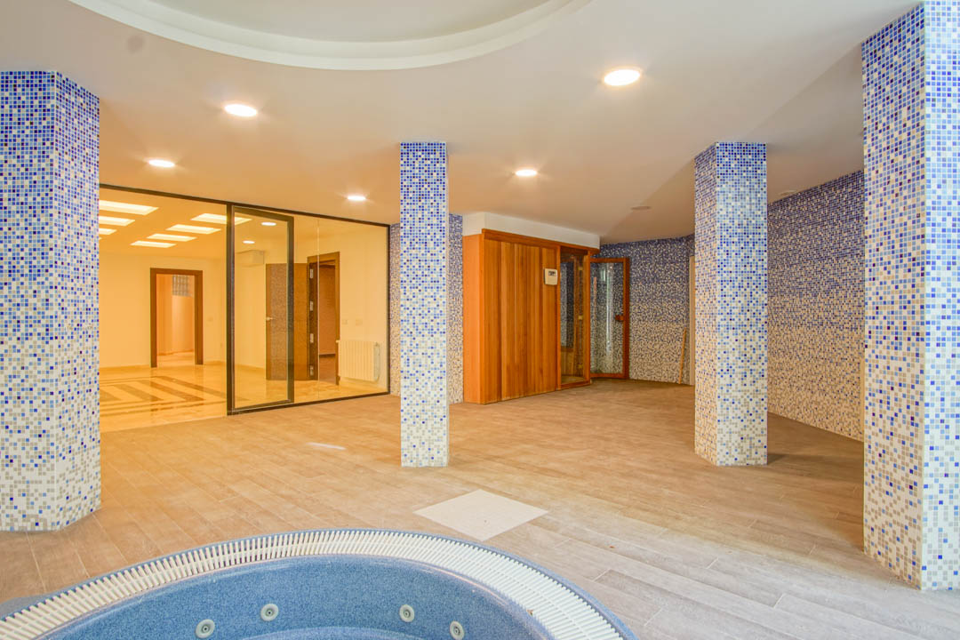 Spa area with a circular jacuzzi, wooden sauna, and blue mosaic-tiled columns. Features recessed ceiling lighting and glass partitions leading to the basement rooms.
