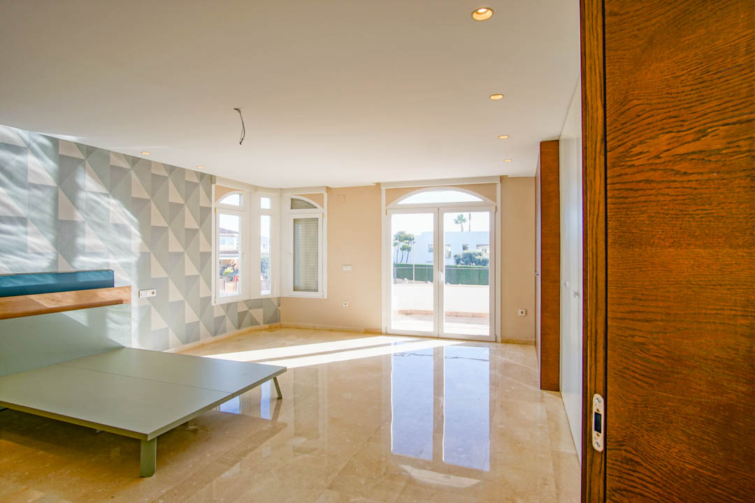 Spacious bedroom featuring polished marble floors, a geometric patterned accent wall, and direct access to a sunny terrace through arched glass doors.