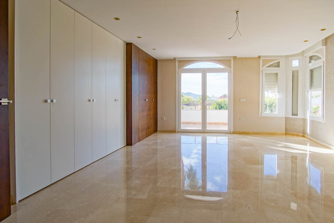 Spacious bedroom featuring polished marble floors, integrated floor-to-ceiling wardrobes in white and wood tones, and arched doors opening to a terrace.