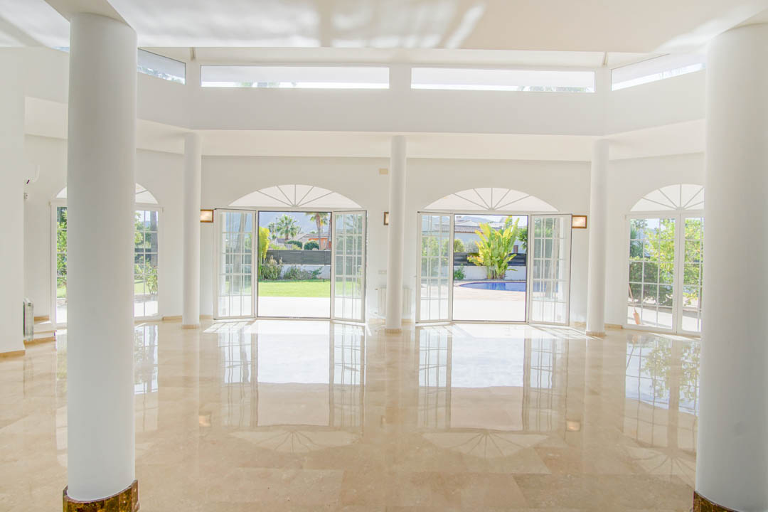 Spacious colonial-style living room featuring circular white columns, polished marble flooring, and arched French doors opening onto the garden and pool area.